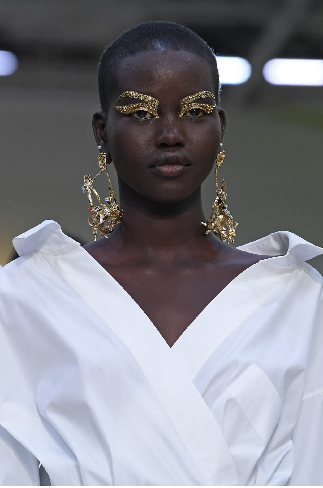 Runway model with sculpted gold-leaf brows and oversized gold earrings